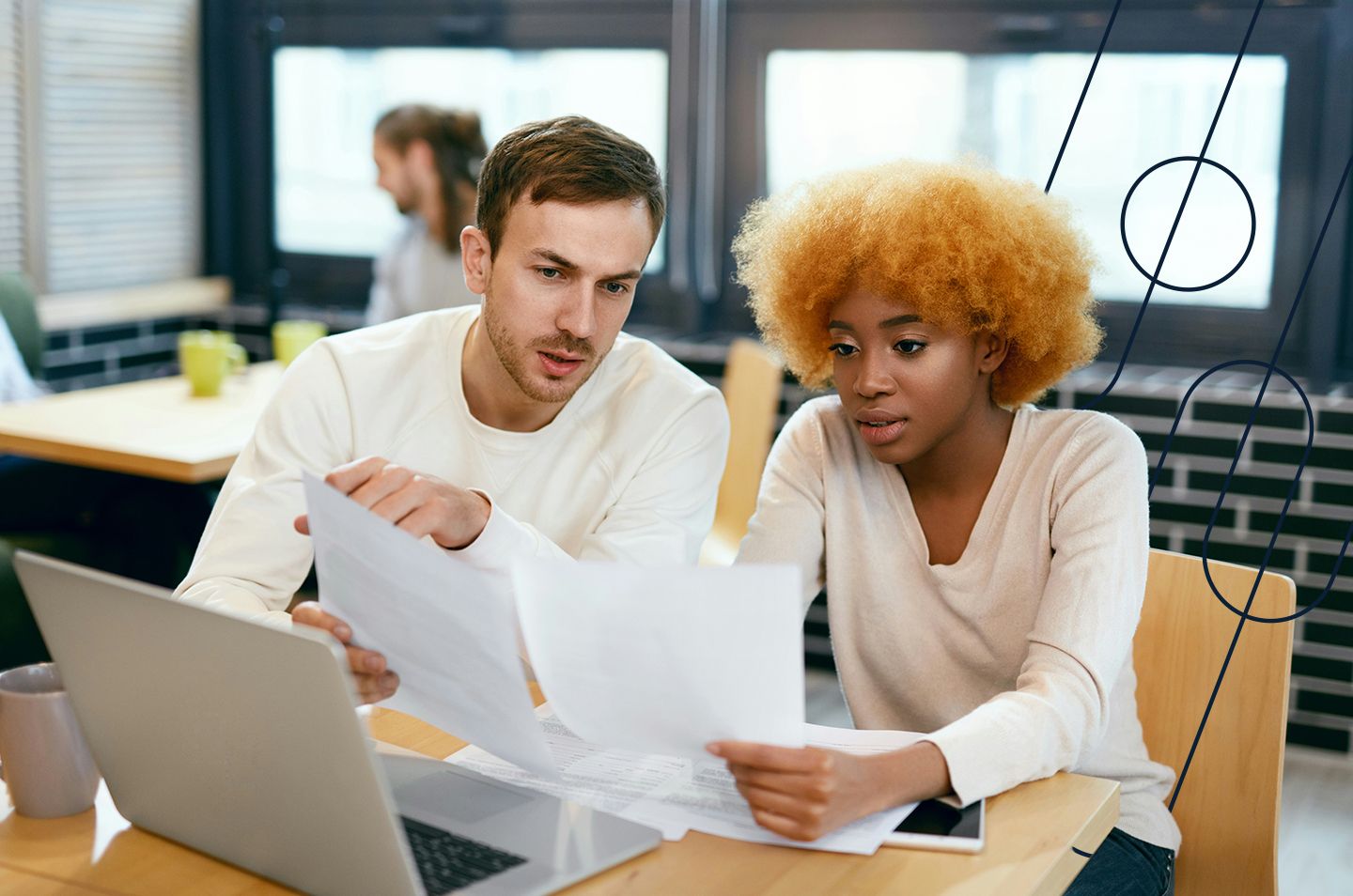 two people sitting at a desk looking at a laptop and paperwork